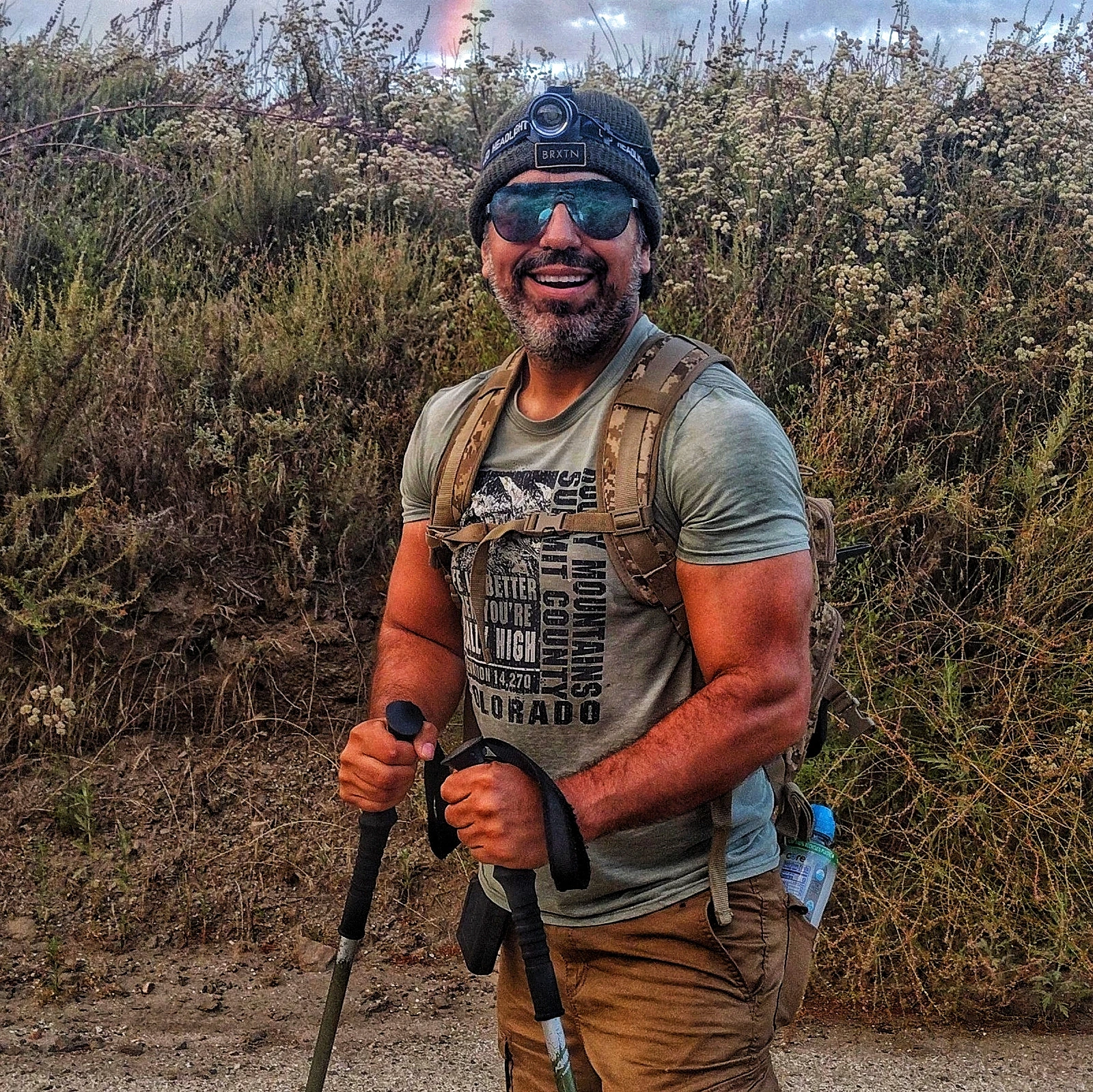 Cazonci Yacata smiling on a mountain trail with trekking poles beneath a rainbow